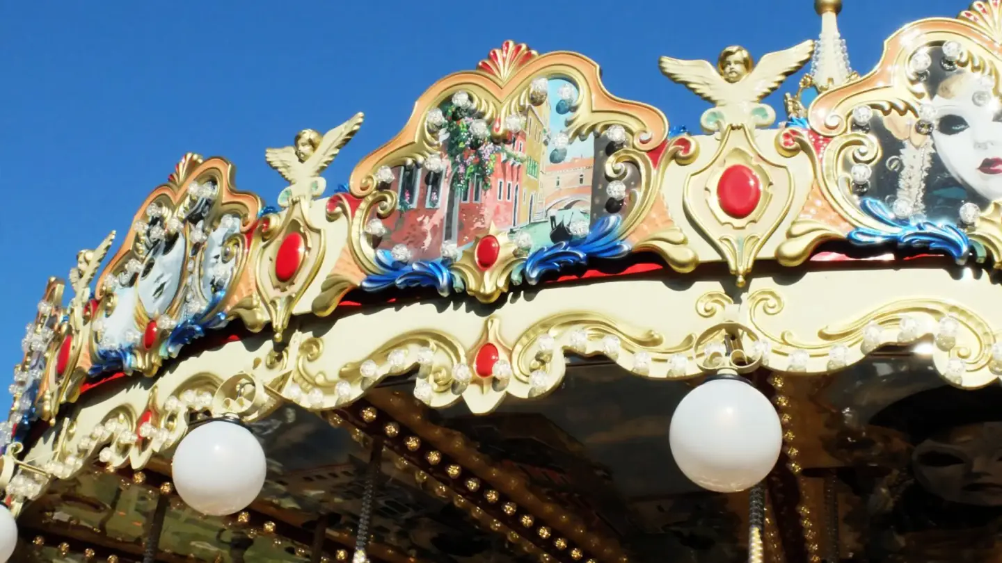 Primer plano de un carrusel veneciano clásico de dos niveles, iluminado con bombillas de luz cálida que resaltan las figuras de caballos y carrozas pintadas a mano en una plaza pública.