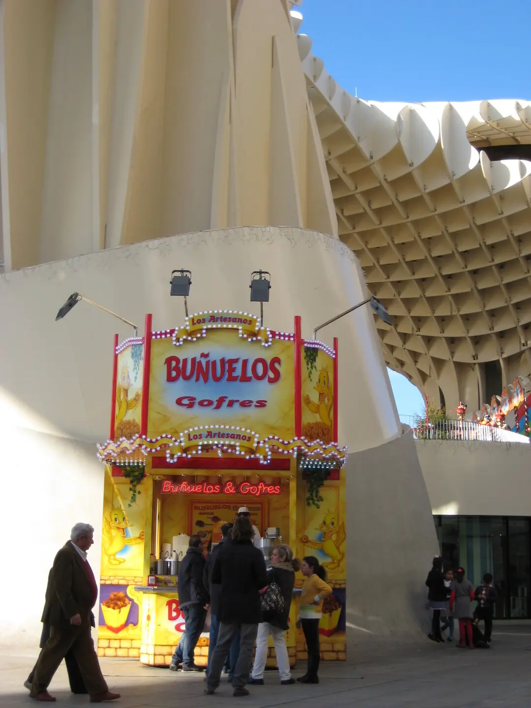 Puesto de buñuelos y gofres de "Los Artesanos" con colores amarillo y rojo, situado en una plaza moderna bajo una estructura arquitectónica blanca.